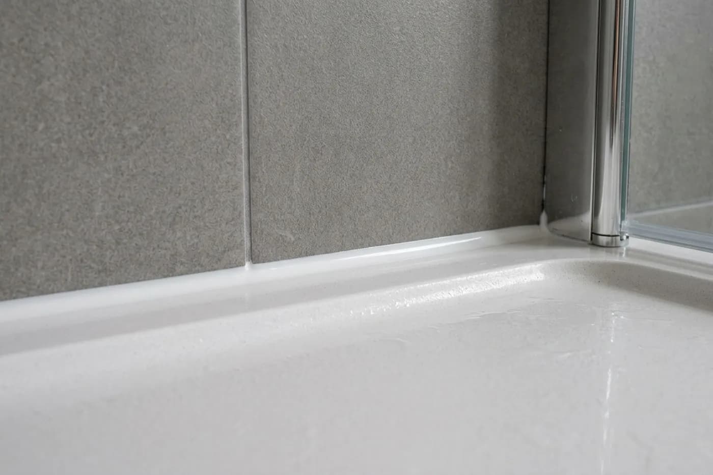 The joint between a white stone resin shower tray and grey wall tiles carrying a fresh white silicone bead tooled to a concave finish