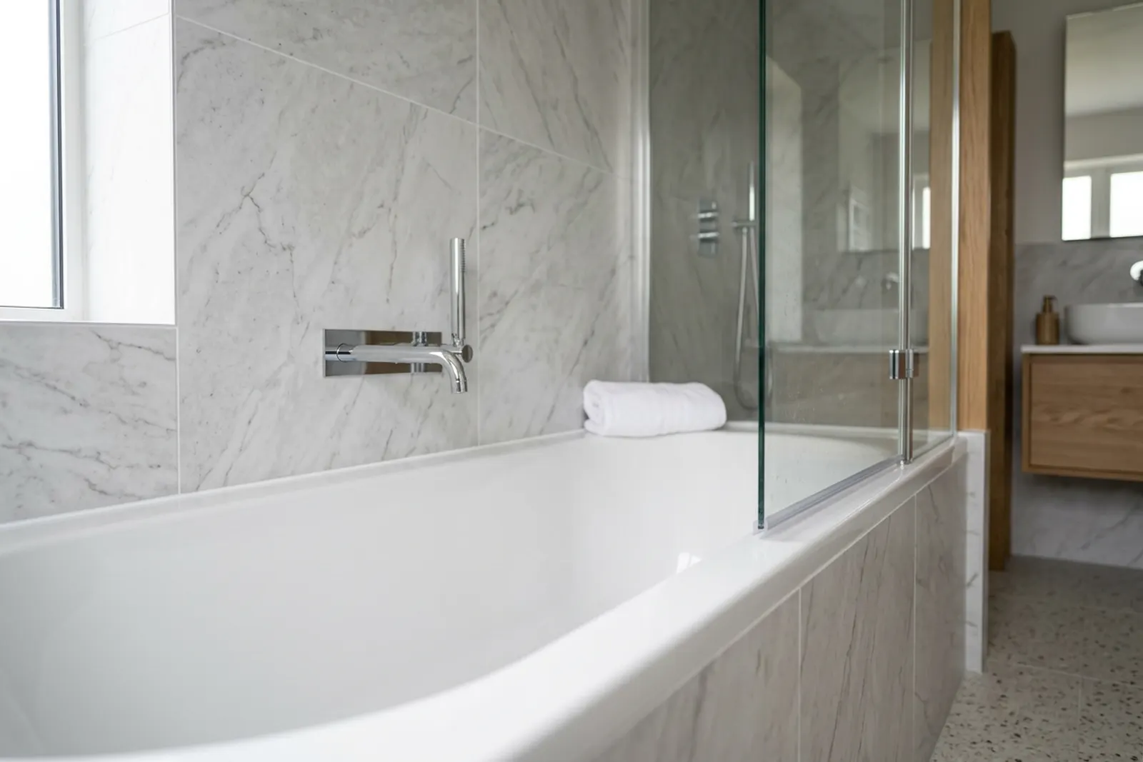 A finished bathroom with pale marble effect wall tiles and a white bath, a clean white silicone bead running along the joint where the bath meets the tile and down the panel, a chrome wall mounted tap, a folded towel and a glass shower screen